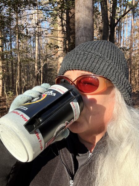 a woman with long white hair wearing a grey stocking hat and sunglasses is drinking matcha from an old travel mug